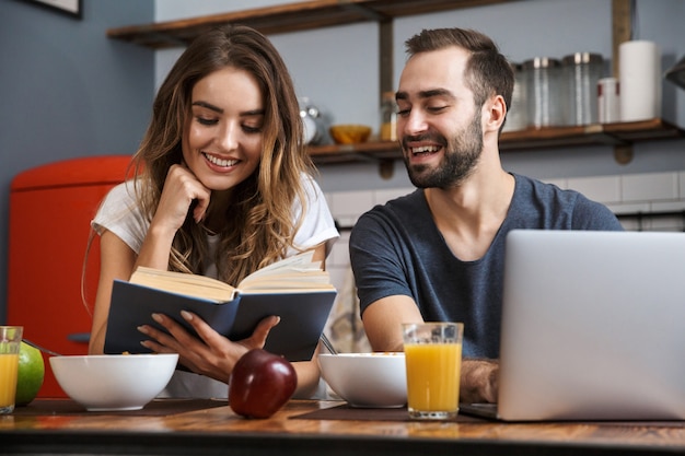 Schönes fröhliches Paar, das Frühstück in der Küche, mit Laptop-Computer, ein Buch lesend hat