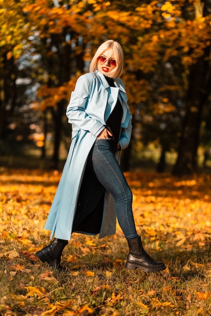 Schönes Geschäftsfrauenmodell mit Sonnenbrille in modischer Kleidung mit Stiefeln spaziert im Park vor dem Hintergrund von orangefarbenem Herbstlaub bei Sonnenuntergang