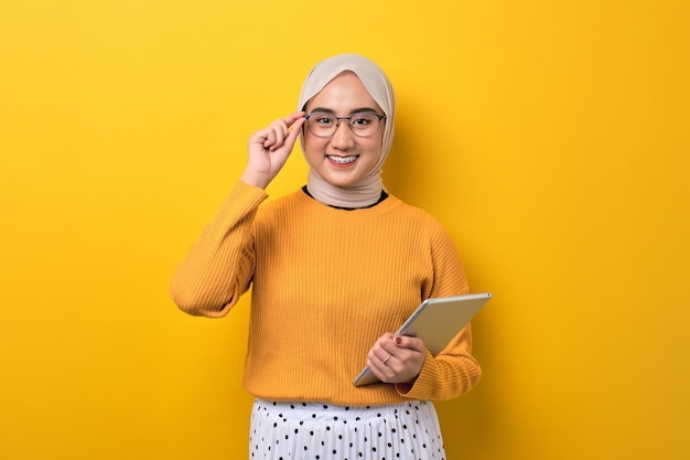 Schönes glückliches asiatisches Mädchen, das Hijab mit digitalem Tablet trägt und in die Kamera lächelt, isoliert auf gelbem Hintergrund