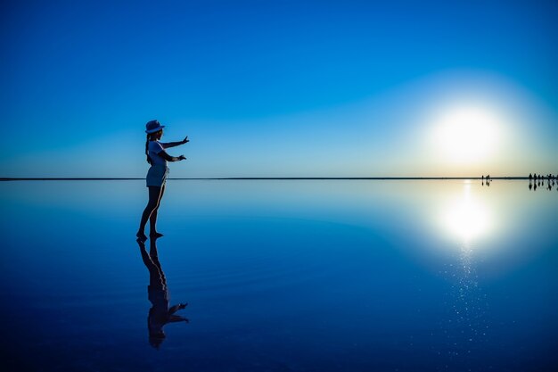 Schönes glückliches junges Mädchen geht und posiert in einer magischen Pose entlang des spiegelrosa Salzsees, genießt die warme Abendsonne und betrachtet den feurigen Sonnenuntergang und ihr Spiegelbild