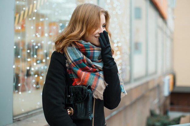 Foto schönes, glückliches, junges model mit überraschenden emotionen, in einem modischen vintage-kartenschal und schwarzem mantel mit einer stilvollen handtasche, geht auf der straße.