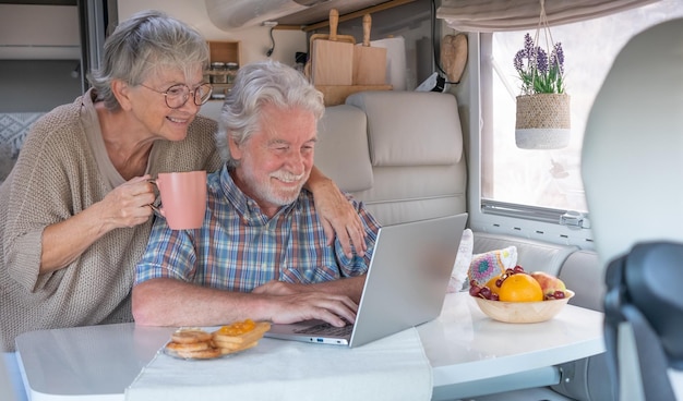 Schönes glückliches kaukasisches Seniorenpaar in der Reiseurlaubsfreizeit, das in einer Essecke eines Wohnmobils sitzt und zusammen frühstückt, während es einen Laptop benutzt