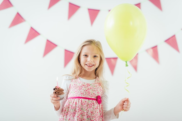 Schönes glückliches kleines Mädchen, das Ballon und kleinen Geburtstagskuchen mit Kerze hält. Blick in die Kamera.