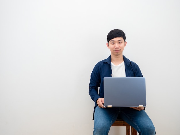 Schönes glückliches Lächeln des asiatischen Mannes, das auf dem Stuhl mit Laptop auf weißem Wandhintergrund sitzt