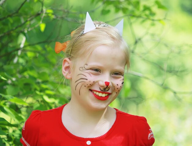 Schönes glückliches lächelndes junges blondes kaukasisches Mädchen mit dem Gesicht, das wie eine Katze auf Sommernaturhintergrund gemalt wird