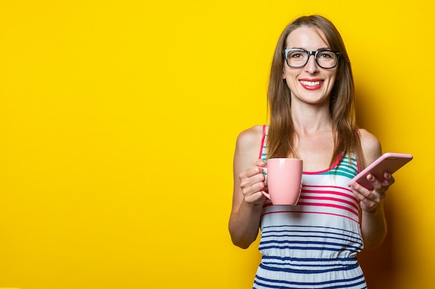 Schönes glückliches lächelndes junges Mädchen, das eine Tasse mit Kaffee und einem Telefon hält