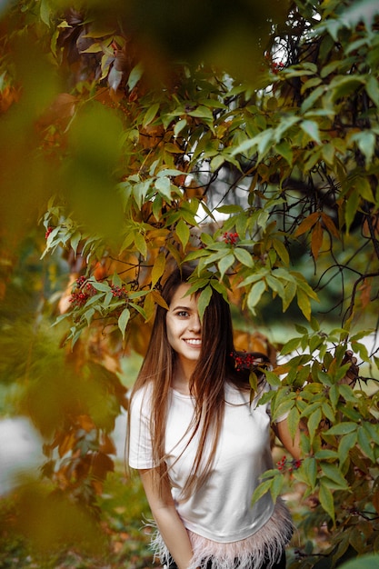 Schönes glückliches lächelndes Mädchen mit langen Haaren, das gegen den Hintergrund des Herbstlaubs im Park aufwirft.