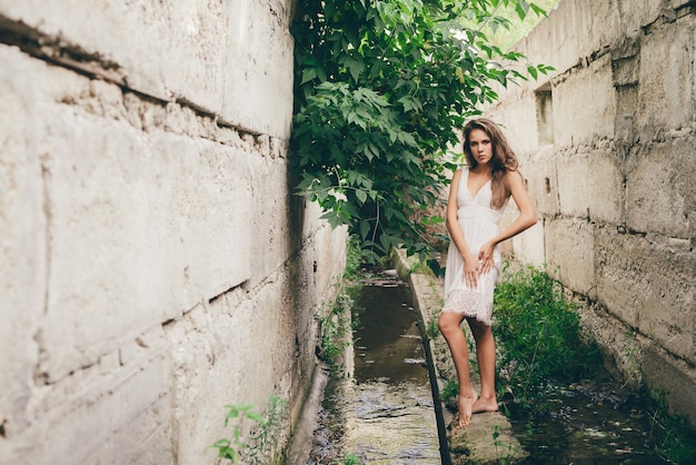 Schönes glückliches Mädchen mit lockigem natürlichem Haar im weißen Kleid nahe grünen Baumblättern