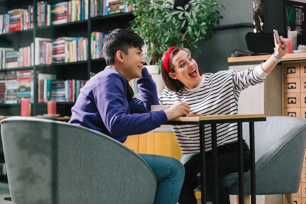 Schönes glückliches Paar, das in einem Café in bequemen Sesseln sitzt und Selfies macht. Bücherregale im Hintergrund