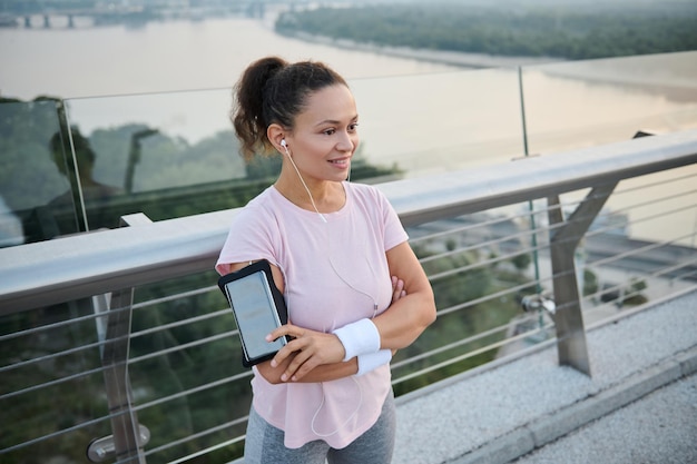 Schönes, glückliches, sportliches Mädchen mit Kopfhörern, die mit verschränkten Armen auf der Stadtbrücke stehen und ein lächelndes, zahniges Lächeln wegschauen. Sportlerin, die sich nach intensivem Cardio-Training im Freien ausruht