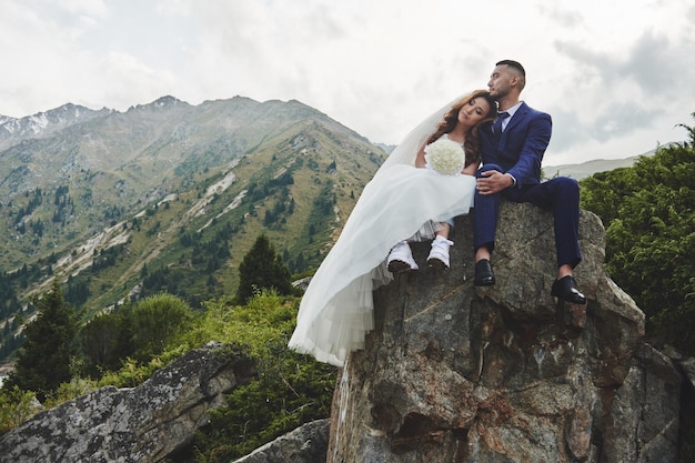 Schönes Hochzeitsfoto auf Bergsee. Glückliches asiatisches verliebtes Paar, Braut im weißen Kleid und Bräutigam im Anzug werden vor dem Hintergrund der kasachischen Landschaft fotografiert