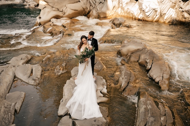 Schönes Hochzeitspaar umarmt zärtlich auf dem Hintergrund eines Gebirgsflusses, langes weißes Kleid der Braut.