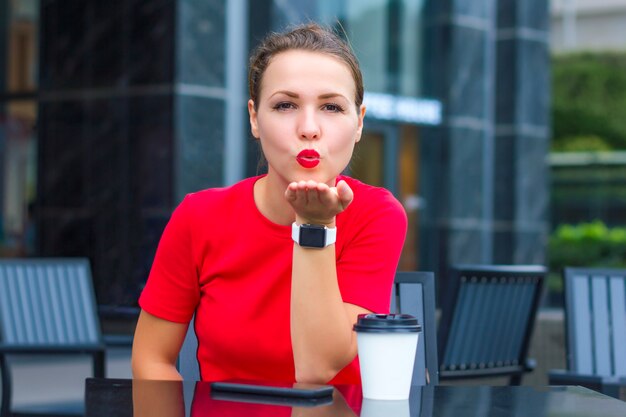 Schönes hübsches Mädchen, junge nette Frau in der roten Sendung, die einen Kuss mit den roten Lippen bläst, direkt in die Kamera schaut, im Café auf der Terrasse mit Tasse Kaffee, Smartphone sitzt. Ich küsse Dich. Dame am Datum