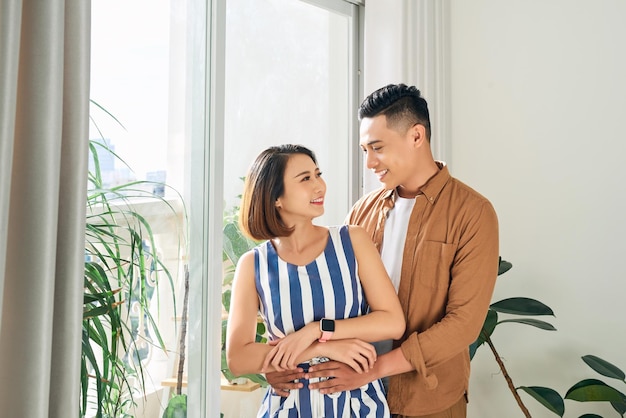 Schönes junges asiatisches verliebtes Paar zu Hause, das am Fenster steht und sich umarmt