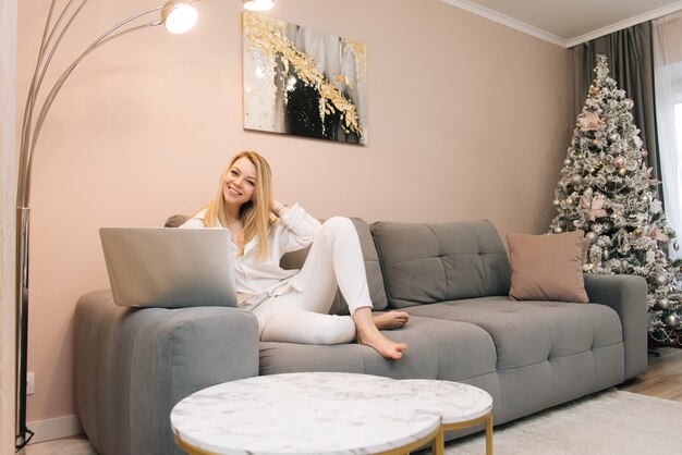 Schönes junges blondes lächelndes Mädchen mit Laptop zu Hause auf der Couch, Fernarbeit von zu Hause, Isolation