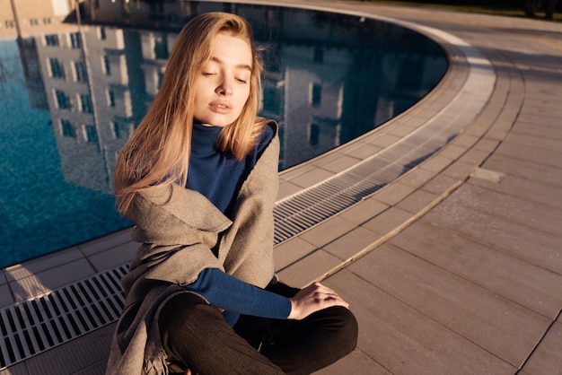 Schönes junges blondes Mädchen im blauen Pullover sitzt am Pool, genießt die Sonne