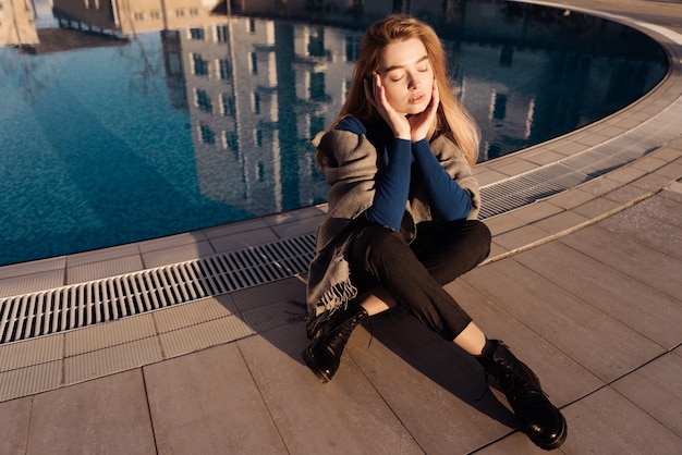 Schönes junges blondes Mädchen in einem blauen Pullover sitzt am Pool in den Strahlen der Frühlingssonne