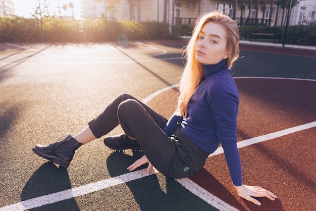 Schönes junges blondes Mädchen in einem blauen Pullover sitzt auf einem Sportplatz in den Morgensonnenstrahlen und posiert