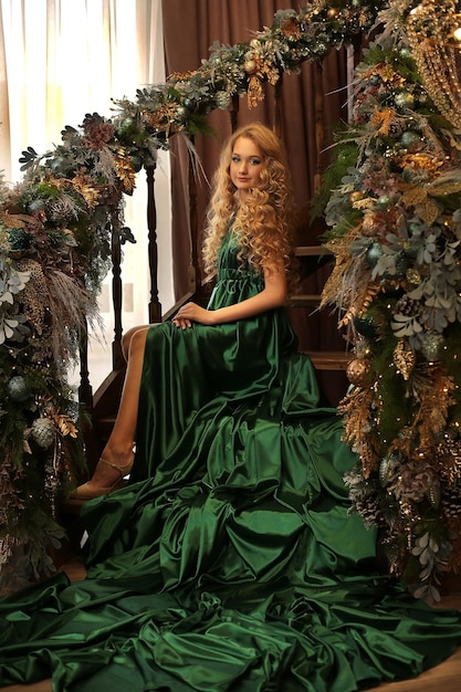 Schönes junges blondes Mädchen in einem schicken grünen Kleid auf der Treppe, das für Weihnachten Neujahr dekoriert ist