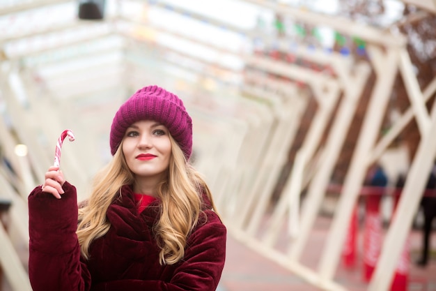 Schönes junges blondes Mädchen in roter Strickmütze posiert mit Zuckerstange auf dem Weihnachtsmarkt