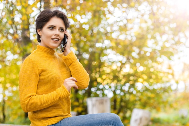 Schönes junges brünettes Mädchen in einem Strickpullover, das im Park mit einem Handy telefoniert