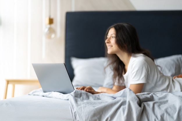 Schönes junges brünettes Mädchen mit einem Laptop, der auf dem Bett liegt. Stilvolles modernes Interieur. Ein gemütlicher Arbeitsplatz. Einkaufen im Internet.