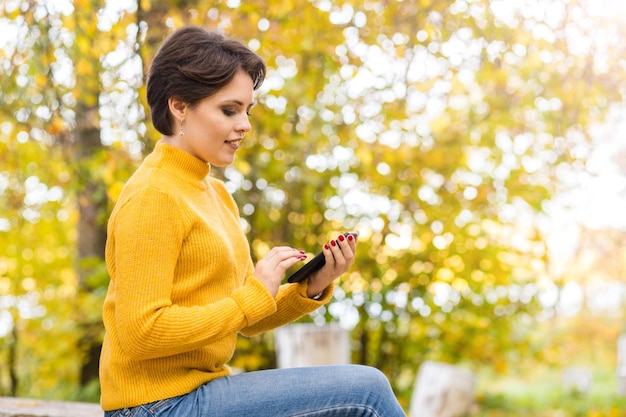 Schönes junges Brunettemädchen, das im Herbstpark mit einem Handy in ihren Händen aufwirft