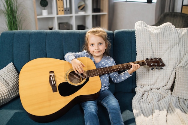 Schönes junges charmantes kleines Mädchen, das lächelt, während es zu Hause klassische Gitarre spielt