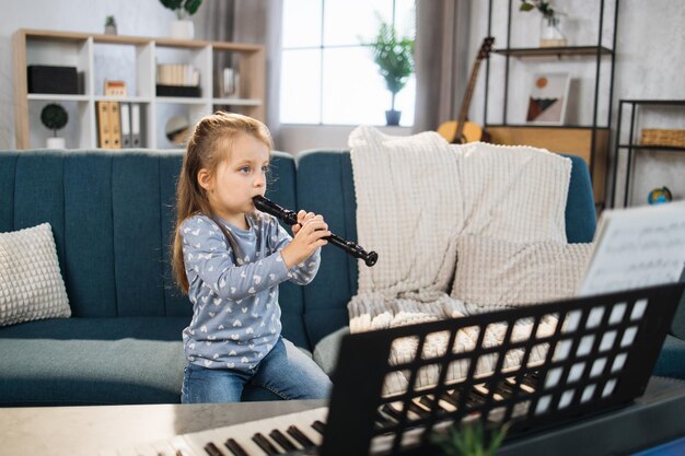Schönes junges charmantes kleines Mädchen lächelt, während es zu Hause klassische Flöte spielt