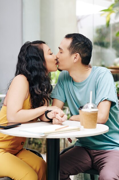 Schönes junges chinesisches paar sitzt am tisch im café im freien, trinkt kaffee und küsst sich