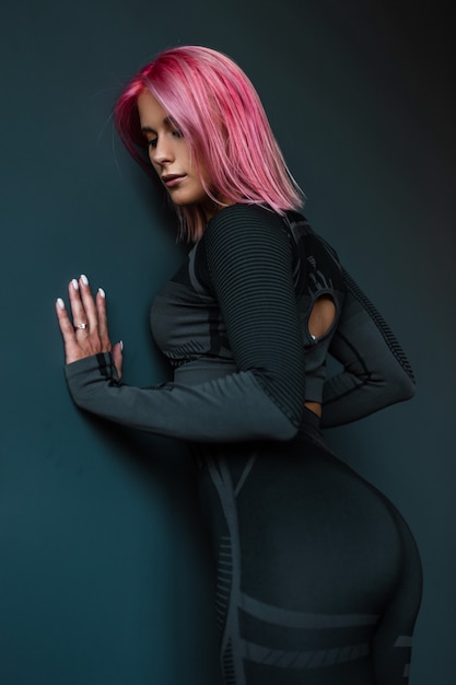 Schönes junges Frauenmodell mit rosa Haaren in der schwarzen Sportkleidung, die nahe einer schwarzen Wand aufwirft