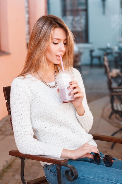 Schönes junges glückliches Frauenmodell in einem modischen Pullover, Jeans und Sonnenbrille sitzt und trinkt einen Milchshake an einem sonnigen Tag