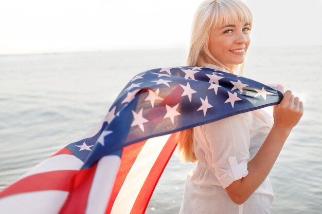 Schönes junges glückliches Mädchen, das amerikanische Flagge nahe Meer hält.