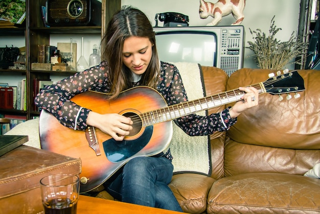 Schönes junges Hippie-Mädchen, das akustische Gitarre spielt, die zu Hause auf einem Sofa sitzt. Retro-Vintage-Farben-Edition