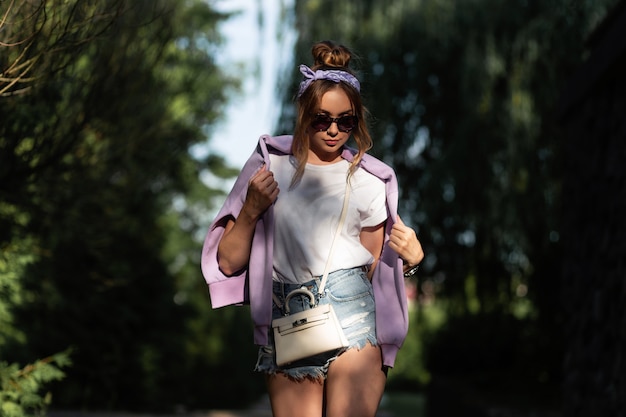 Schönes junges Hipster-Mädchen mit Vintage-Bandana und Sonnenbrille in modischen Denim-Shorts und weißem T-Shirt mit violettem Kapuzenpulli Spaziergang in der Natur