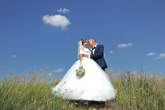 Schönes junges Hochzeitspaar, das oben auf einem Hügel auf einem Hintergrund des blauen Himmels und der Wolken steht.