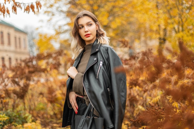 Schönes junges hübsches Frauenmodell in modischer Freizeitkleidung mit einer Lederjacke und einem Pullover mit einer Tasche geht im Herbstpark spazieren