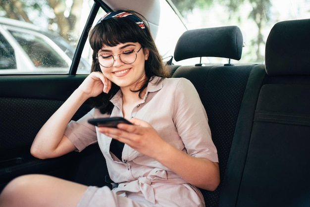 Schönes junges hübsches Mädchen benutzt ein Smartphone und lächelt, während es auf dem Rücksitz im Auto sitzt