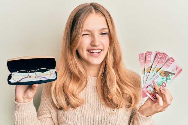 Schönes junges kaukasisches Mädchen, das eine Brille und Neuseeland-Dollar hält und mit einem fröhlichen und glücklichen Gesicht in die Kamera schaut