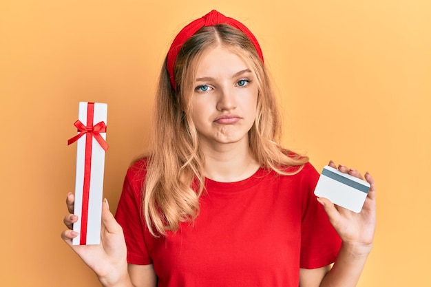 Schönes junges kaukasisches Mädchen, das Geschenk und Kreditkarte deprimiert hält und sich Sorgen um Not macht und wütend und ängstlich traurigen Ausdruck weint