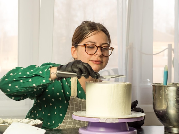 Schönes junges Konditormädchen macht mit einem Kochspatel einen Kuchen mit weißer Sahne