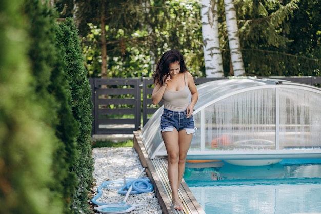 Schönes junges Mädchen, das auf dem Hintergrund des Pools steht und telefoniert, sie ist glücklich. Sie trägt ein stilvolles weißes T-Shirt mit einem Aufdruck von Palmen, blauem Rucksack und blauer Brille