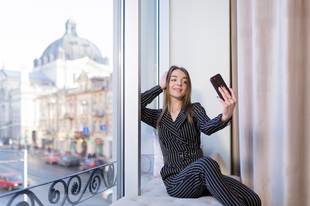 schönes junges Mädchen, das ein Selfie mit Telefon auf der Fensterbank des Hotels gegen die Stadt nimmt