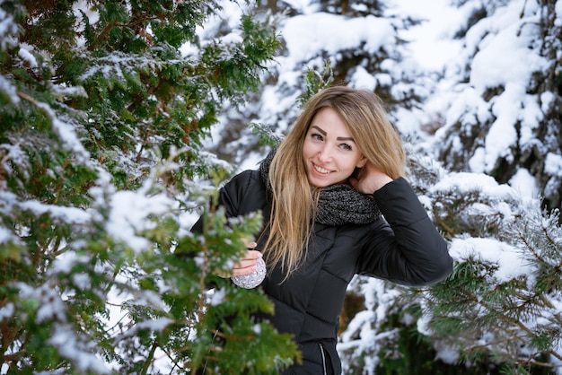 Schönes junges Mädchen, das im Winterwald in den Bäumen steht und Weihnachtskugel hält, lächelt und lacht ...