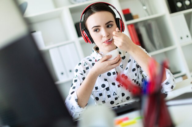 Schönes junges Mädchen, das in den Kopfhörern am Schreibtisch im Büro sitzt, Joghurt isst und Monitor betrachtet.