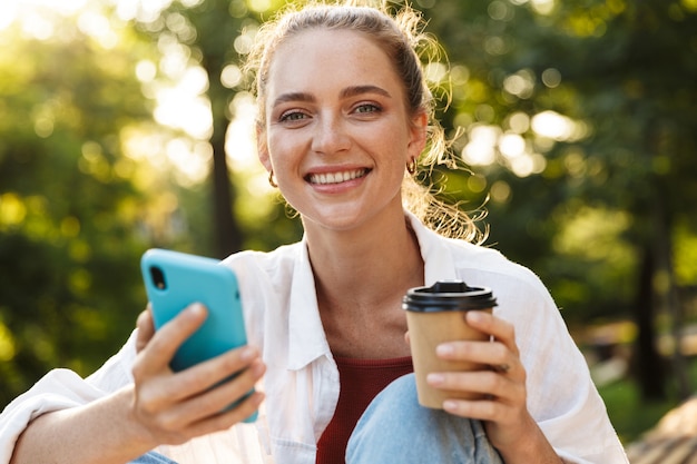 Schönes junges Mädchen, das lässig gekleidet auf einer Bank im Stadtpark sitzt und Kaffee trinkt, während es das Handy benutzt