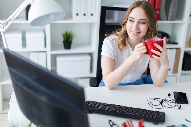 Schönes junges Mädchen, das mit Computer im Büro arbeitet. Das Mädchen hat eine rote Tasse in den Händen.