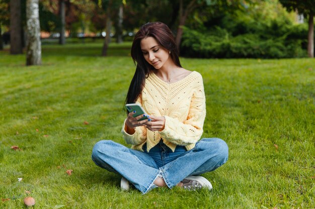 Schönes junges Mädchen, das mit einem Smartphone ein Selfie-Foto im Park macht. Mädchen mit einem Telefon, das auf dem Gras im Park sitzt.