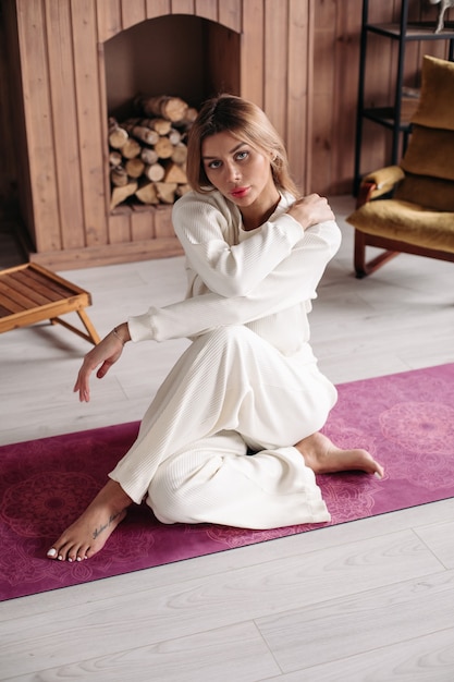 Schönes junges Mädchen, das natürliche organische umweltfreundliche weiße Baumwollsportkleidung trägt und auf Matte in gemütlichem Wohninnenraum sitzt. Foto in hoher Qualität