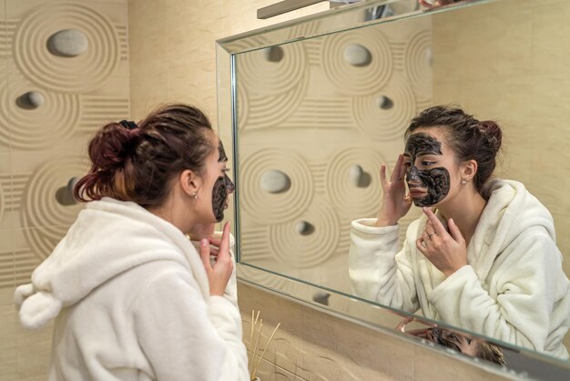 Schönes junges Mädchen im Badezimmer legt eine schwarze Tonmaske und schaut in den Spiegel Das Konzept einer kosmetischen Maske auf dem Gesicht